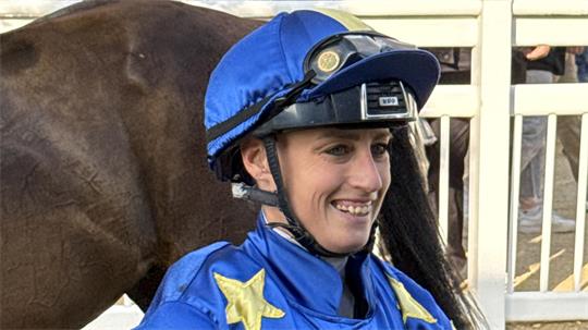 Jockey mit blauem Helm und blauer Jacke mit gelben Sternen lächelt neben einem braunen Pferd.