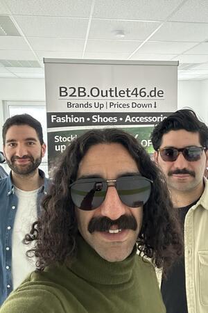 Drei Personen vor einem Werbebanner mit der Aufschrift 'B2B.Outlet46.de Brands Up | Prices Down Fashion • Shoes • Accessori'