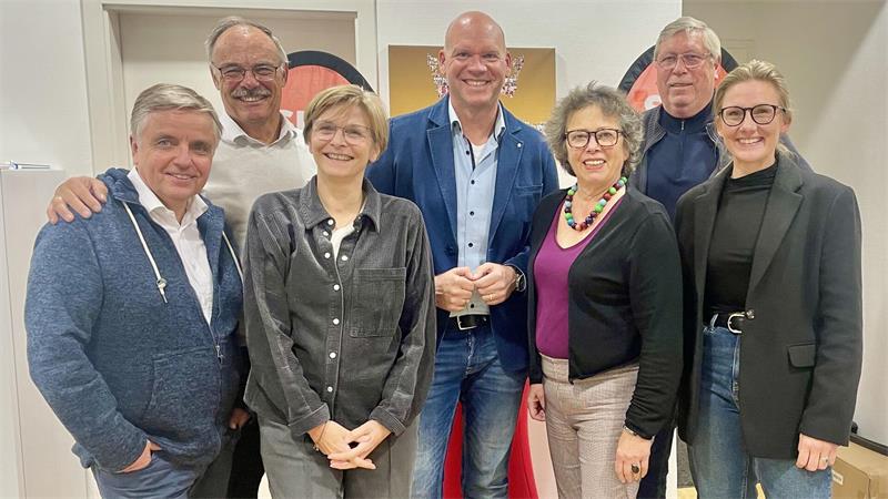 Gruppe von sieben Personen unterschiedlichen Alters steht eng beieinander und lächelt in einem Innenraum
