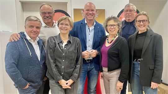 Gruppe von sieben Personen unterschiedlichen Alters steht eng beieinander und lächelt in einem Innenraum