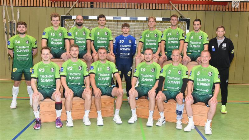Mannschaftsbild einer Handballmannschaft in grünen Trikots und einem Spieler in blauem Torwarttrikot in einer Sporthalle vor einem Handballtor
