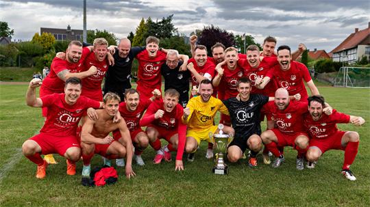 Mannschaft in roten Trikots jubelt auf Fußballfeld um Pokal auf dem Boden