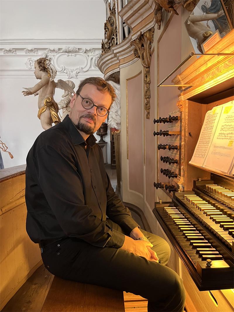 Ein schwarz gekleideter Mann mit Bart sitzt an einer beleuchteten Orgel, hinter ihm eine Putte.