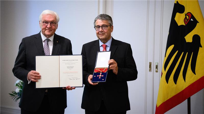 Zwei Personen stehen in einem Raum und halten eine Urkunde sowie ein Bundesverdienstkreuz in die Kamera