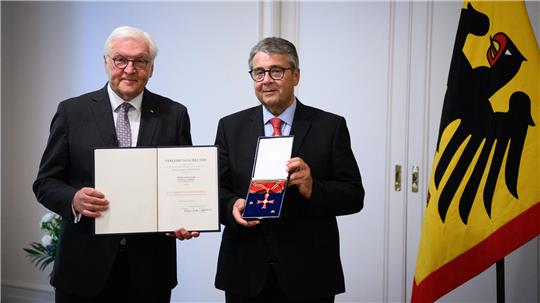 Zwei Personen stehen in einem Raum und halten eine Urkunde sowie ein Bundesverdienstkreuz in die Kamera