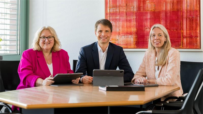 Zwei Frauen und ein Mann sitzen an einem Tisch und lachen in die Kamera.