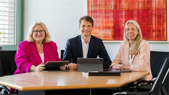 Zwei Frauen und ein Mann sitzen an einem Tisch und lachen in die Kamera.