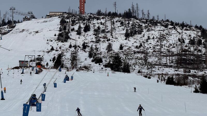 Schneebedeckter Hang mit Skilift, mehreren Skifahrern und kahlen Bäumen unter bewölktem Himmel.