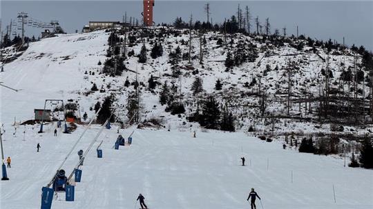 Schneebedeckter Hang mit Skilift, mehreren Skifahrern und kahlen Bäumen unter bewölktem Himmel.