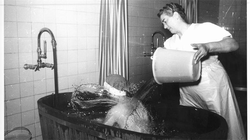 Ein Mann sitzt in einer Badewanne, eine Frau gießt ihm Wasser über den Rücken.
