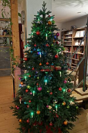 Ein künstlicher Weihnachtsbaum, geschmückt. Einmal um ihn herum fährt auf mittlerer Höhe eine Modelleisenbahn.