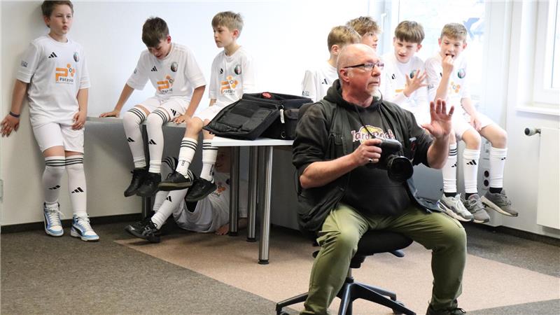 Gruppe von Jungen in weißen Sporttrikots sitzt an einer Wand und unter einem Tisch, ein Mann mit Kamera sitzt auf einem Stuhl und hebt die Hand