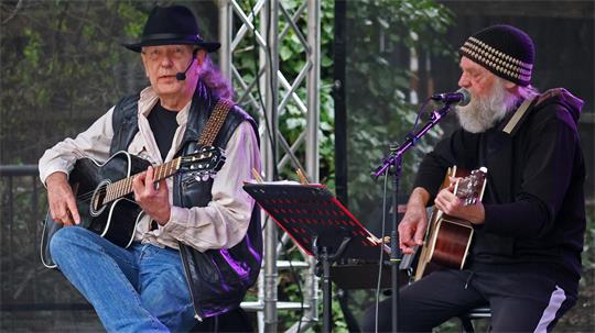 Zwei ältere Männer mit Gitarren sitzen auf einer Bühne und singen in Mikrofone