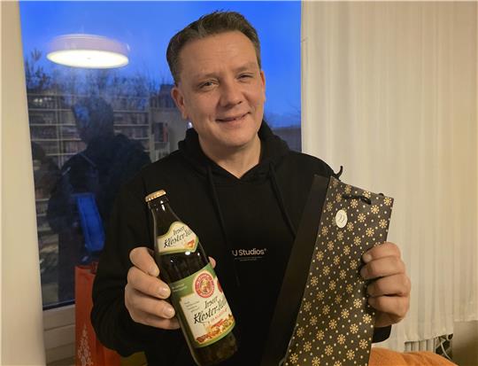 Mann in schwarzem Kapuzenpullover hält eine Flasche Klosterbier und eine Geschenkverpackung mit goldfarbenem Muster vor einem Fenster mit Blick auf den Abendhimmel.