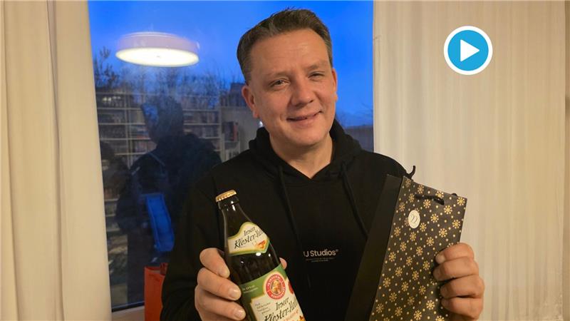 Mann in schwarzem Kapuzenpullover hält eine Flasche Klosterbier und eine Geschenkverpackung mit goldfarbenem Muster vor einem Fenster mit Blick auf den Abendhimmel.