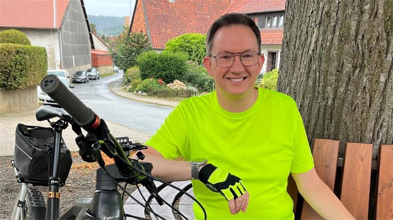 Ein Mann sitzt auf einer Bank und lächelt in die Kamera. Er trägt Sportkleidung. Neben ihm steht ein Fahrrad.