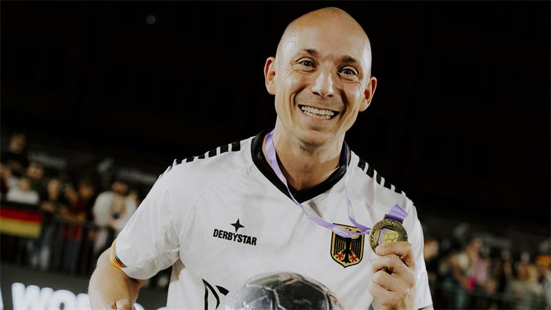 Mann in weißem Trikot mit Medaille hält großen Pokal auf Spielfeld mit jubelndem Publikum im Hintergrund