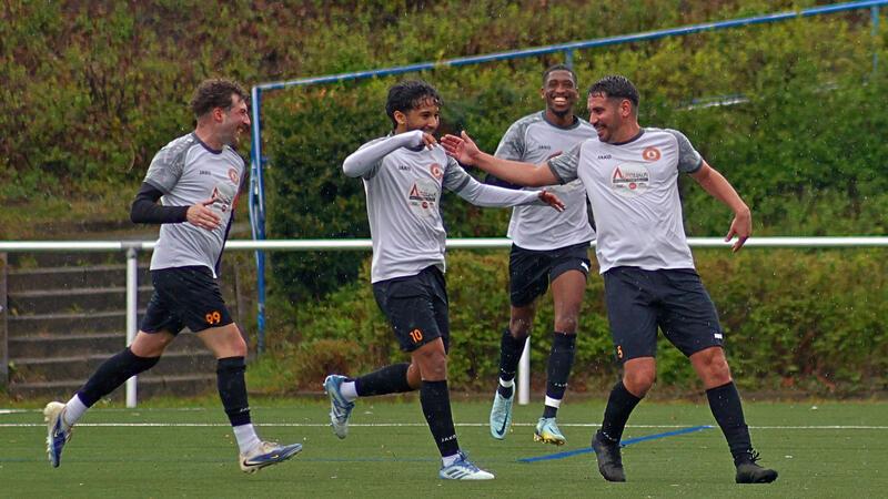 Vier Fußballspieler in weißen Trikots und schwarzen Hosen auf einem Spielfeld feiern mit ausgestreckten Armen