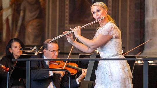 Eine Flötistin in weißem Kleid spielt vor zwei Geigern in einem Konzertsaal.