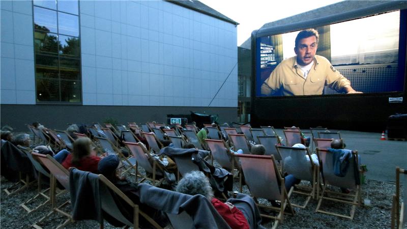 Menschen sitzen in Liegestühlen vor einer großen Leinwand im Freiluftkino
