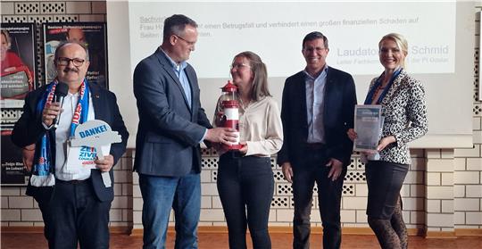 Fünf Personen stehen nebeneinander, zwei Männer und drei Frauen, eine Frau hält eine Flasche, eine andere eine Urkunde, im Hintergrund eine Projektionswand mit Text.