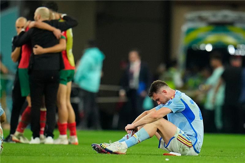Spanien tröstet sich nach Abflug: „Junge Truppe mit Zukunft” Spaniens Aymeric Laporte sitzt nach der Niederlage auf dem Spielfeld, während Marokkos Spieler im Hintergrund über ihren Sieg im Elfmeterschiessen jubeln.