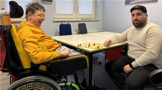 Ein Mann im Rollstuhl und ein anderer Mann sitzen sich an einem Tisch gegenüber und spielen Mensch-ärgere-dich-nicht.