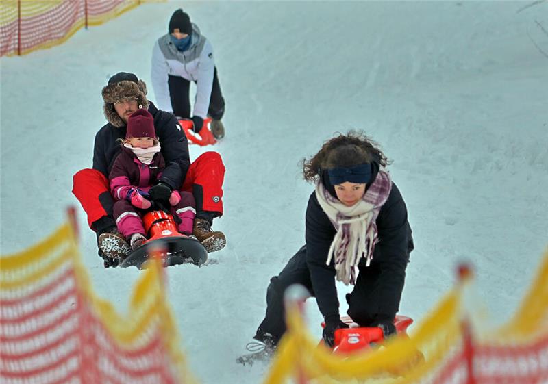 Mehrere Personen rodeln auf einer schneebedeckten Bahn, umgeben von gelb-rotem Sicherheitsnetz.