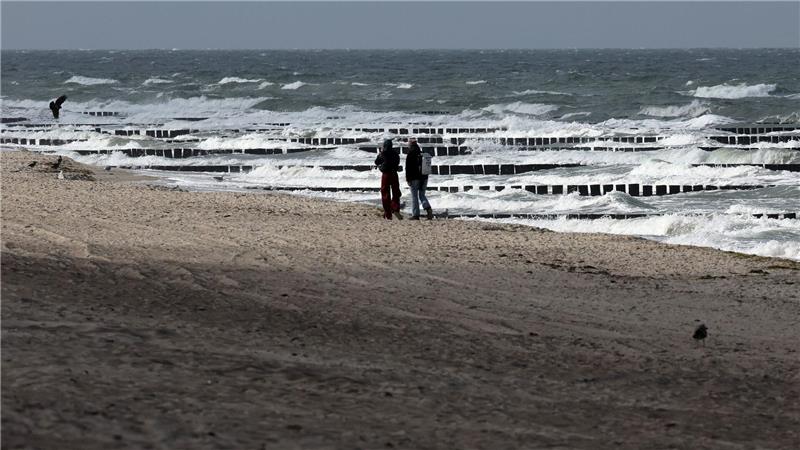Spaziergänger sind am Ostseestrand bei Graal-Müritz unterwegs, wobei nur ab und zu etwas Sonne durch die grauen Wolken am Himmel fällt.