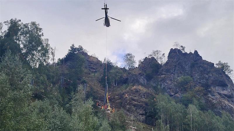 Ein Hubschrauber transportiert Last an Seil über bewaldeten Berghang mit Felsen.