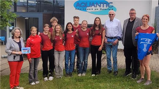 Gruppe von elf Personen vor einem Gebäude mit dem Schriftzug 'AQUANTIC Schwimmsport am Osterfeld', einige tragen rote Shirts, eine Person hält ein blaues T-Shirt mit weißem Aufdruck