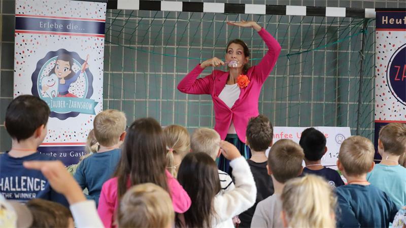 Frau in pinkem Oberteil zeigt Kindern mit erhobenen Armen eine Bewegung in einem Klassenraum mit Postern an der Wand