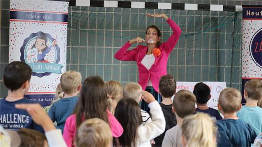 Frau in pinkem Oberteil zeigt Kindern mit erhobenen Armen eine Bewegung in einem Klassenraum mit Postern an der Wand