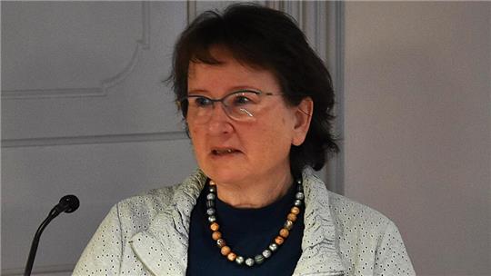 Frau mit kurzen dunklen Haaren trägt eine Perlenkette und einen hellen Pullover über einem dunklen Oberteil