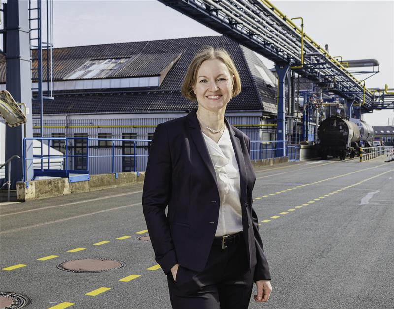 Frau in dunklem Anzug mit weißen Hemd steht auf einer Straße vor einem Industriegebäude mit Rohrleitungen und einem Tankwagen