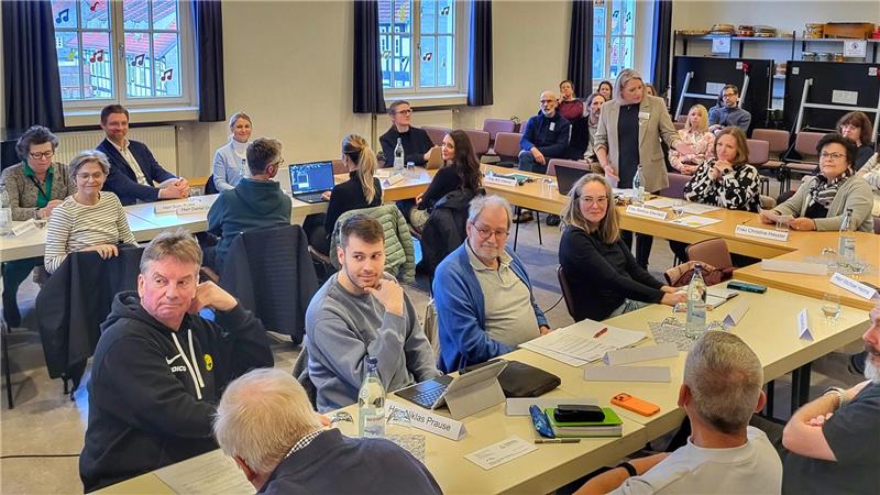 Mehrere Personen sitzen an Tischen in einem hellen Raum und blicken in Richtung eines Sprechers oder einer Präsentation