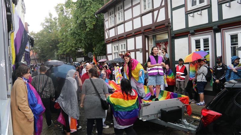 Menschenmenge auf einer Straße bei Regen mit Regenbogenfahnen und -umhängen vor einem Fachwerkhaus.