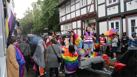 Menschenmenge auf einer Straße bei Regen mit Regenbogenfahnen und -umhängen vor einem Fachwerkhaus.
