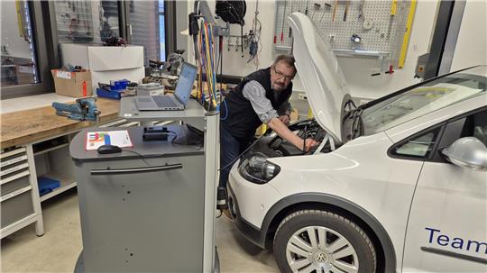 Person arbeitet mit geöffnetem Motorraum an einem weißen Auto in einer Werkstatt mit Werkzeugwand und Werkbänken.