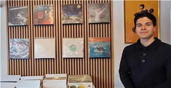 Person steht in einem Raum mit Holzdecke und Wandverkleidung aus Holzlatten vor einer Wand mit sechs Schallplatten-Covern und mehreren Kisten mit Vinyl-Schallplatten auf einem Tisch.