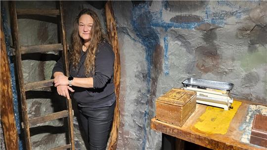 Frau mit langen, lockigen Haaren, schwarzem Pullover und Lederhose lehnt an einer Holzleiter in einem Raum mit bemalter Steinwand und Holztisch.