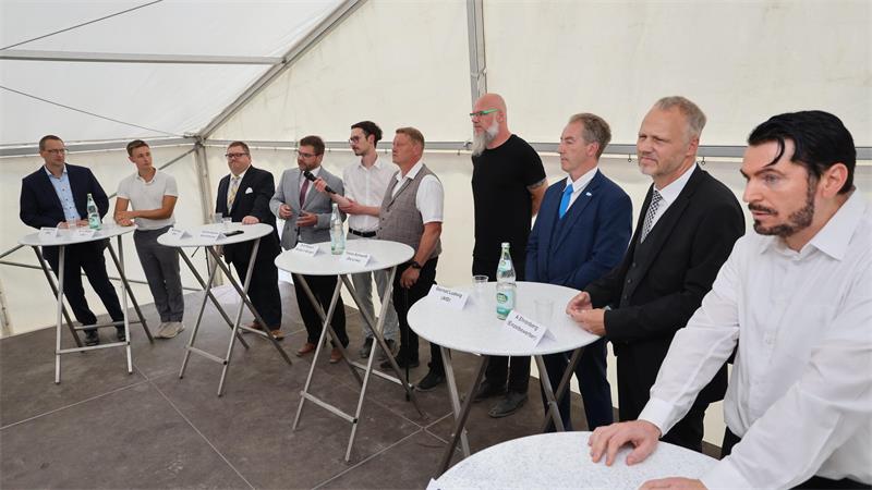 Zehn Männer stehen an Stehtischen auf einer Bühne.