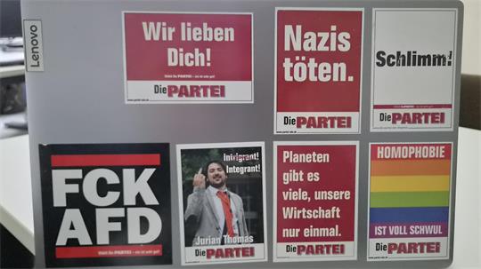 Laptopdeckel mit mehreren rechteckigen Aufklebern der Partei Die PARTEI, darunter Slogans wie 'Wir lieben Dich', 'Nazis töten', 'Schlimm!', 'FCK AFD', 'Intrigant! Integrant!', 'Planeten gibt es viele, unsere Wirtschaft nur einmal' und ein Regenbogen-Aufkleber mit 'MOMOPHOBIE IST VOLL SCHWUL'.