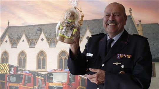 Stellvertender Stadtbrandmeister a.D. Frank Slotta steht stolz, mit einem Abschiedsgeschenk in der Hand und in Uniform vor einer Leinwand mit den Einsatzfahrzeugen der Feuerwehr Goslar.