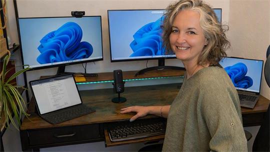 Das Foto zeigt Stephanie Bähr am Arbeitsplatz. Vor ihr stehen zwei Monitore, rechts und links hat sie Laptops, dazwischen ein Mikrofon für SPrachaufnahme.