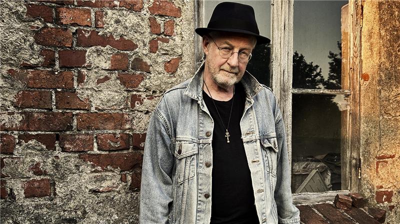 Ein Mann mit Hut und Jeansjacke steht vor einer alten etwas mitgenommenen Mauer mit Fenster.