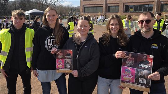 Fünf Menschen stehen auf einem Schulhof, einer trägt eine gelbe Warnweste, vier halten Plakate mit Musik- und Veranstaltungsmotiven