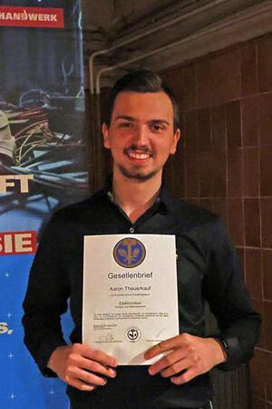 Warum sich Aaron Theuerkauf früh fürs Handwerk entschieden hat Person hält einen Gesellenbrief vor einem Plakat