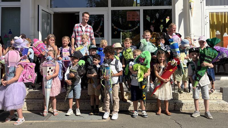 Gruppe von Kindern mit Schultüten und Rucksäcken steht vor einem Schulgebäude, begleitet von zwei Erwachsenen