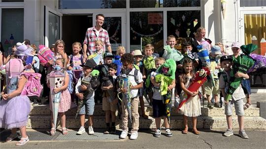Gruppe von Kindern mit Schultüten und Rucksäcken steht vor einem Schulgebäude, begleitet von zwei Erwachsenen